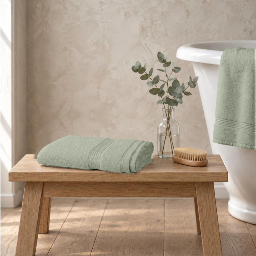 Sage green Egyptian cotton luxury bath sheet, 650gsm, folded on an oak bench beside a roll-top bath