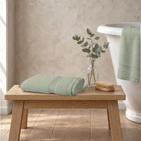 Sage green Egyptian cotton luxury bath sheet, 650gsm, folded on an oak bench beside a roll-top bath