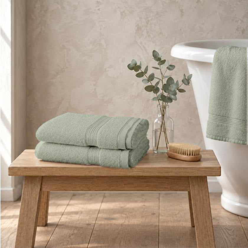 Set of two sage green Egyptian cotton luxury bath sheets, 650gsm, stacked on an oak bench beside a roll-top bath