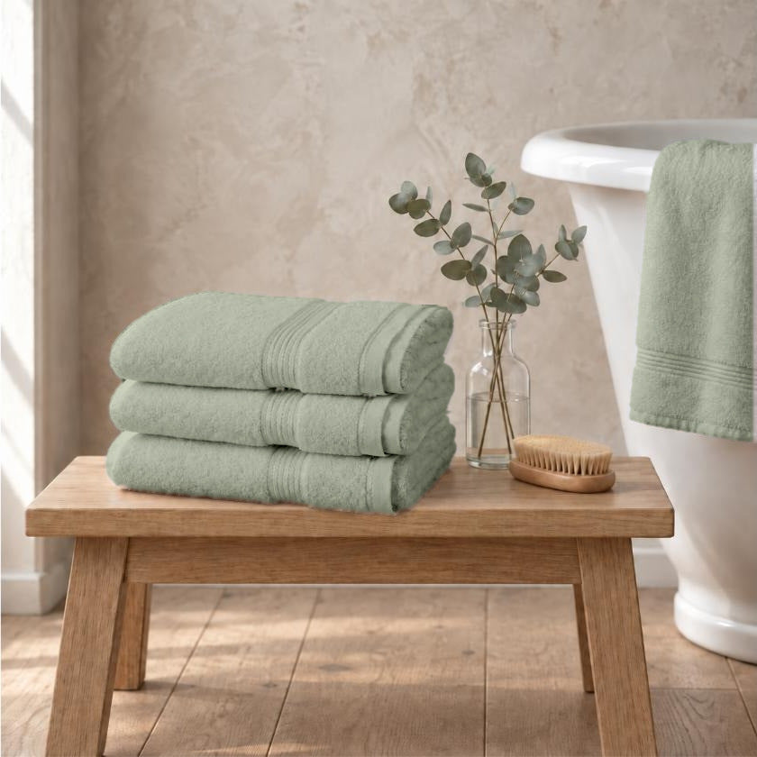 Set of three sage green Egyptian cotton luxury bath sheets, 650gsm, stacked on an oak bench beside a roll-top bath