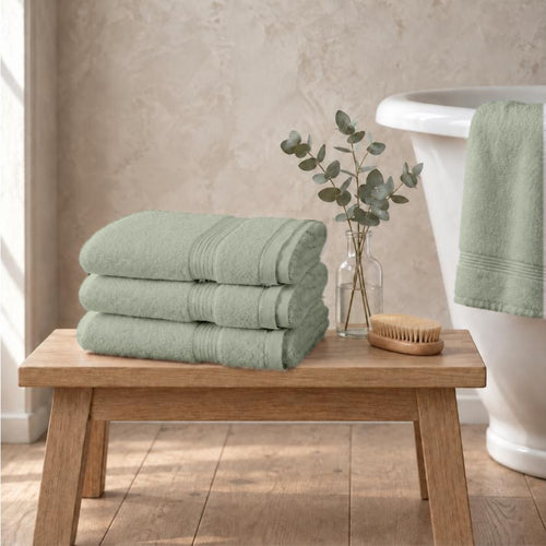 Set of three sage green Egyptian cotton luxury bath sheets, 650gsm, stacked on an oak bench beside a roll-top bath